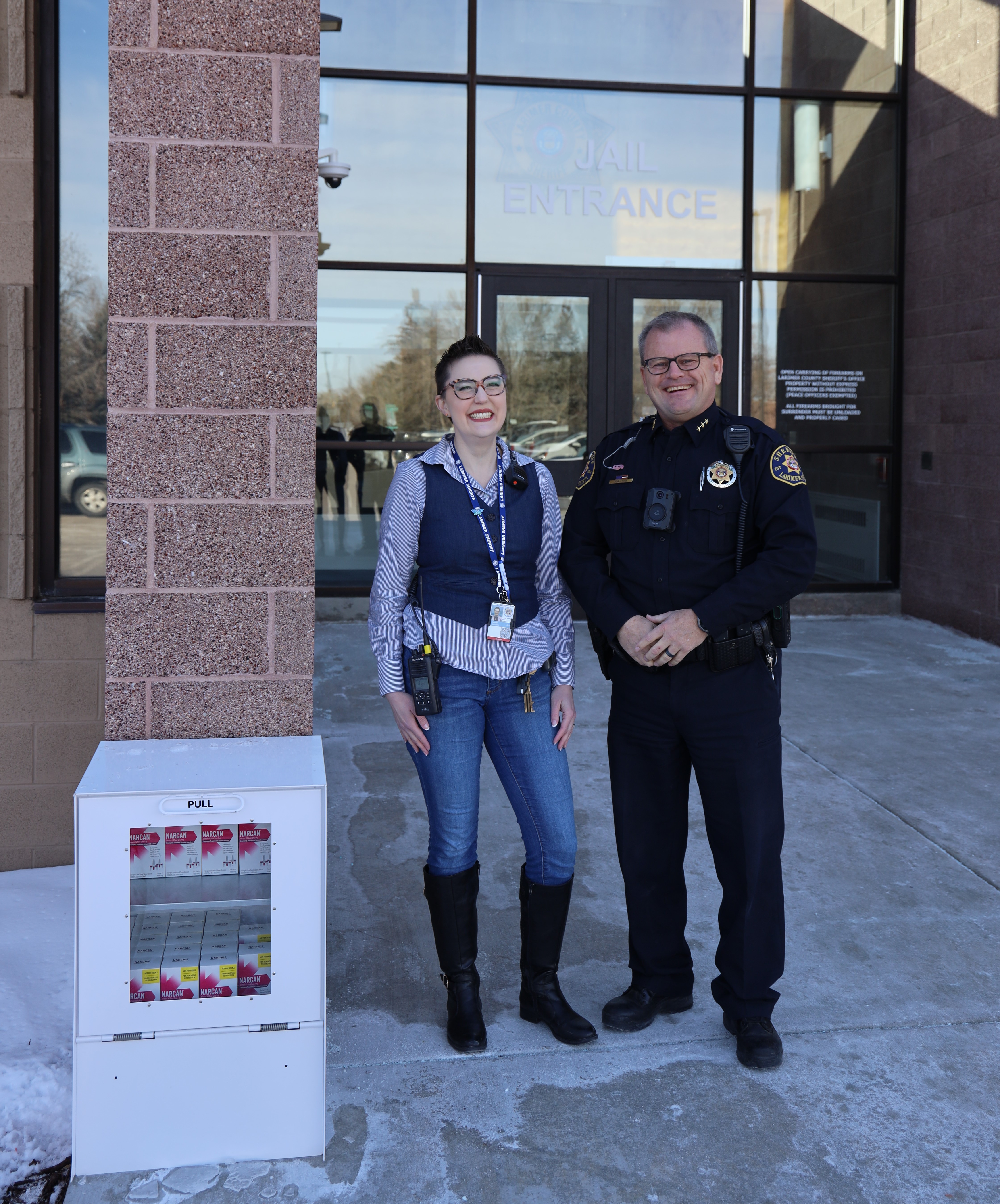 Image 1: Stone Soup Program Coordinator Janelle Goodwin and Sheriff John Feyen stand next to a new Narcan kiosk outside the Larimer County Jail