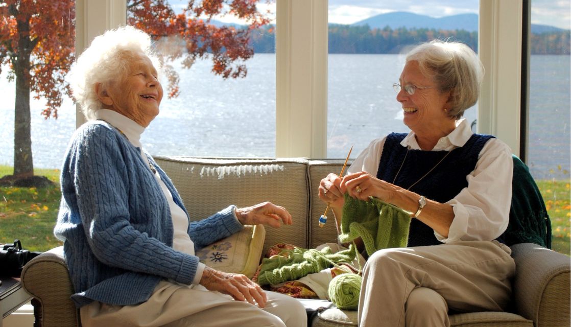 Two older adults laughing and knitting in front of a window showing a sunny mountain lake in fall.