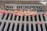 A storm drain grate with dry leaves scattered on top. A metal sign behind the grate reads ‘NO DUMPING – DRAINS TO CREEK
