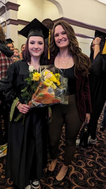 A woman in a graduation cap and gown stands next to another woman, who is smiling.