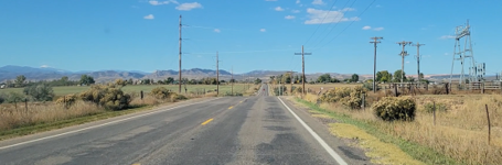 County Road 70 stretching straight through a rural landscape toward distant mountains under a clear blue sky, lined with utility poles, dry brush, and fields.