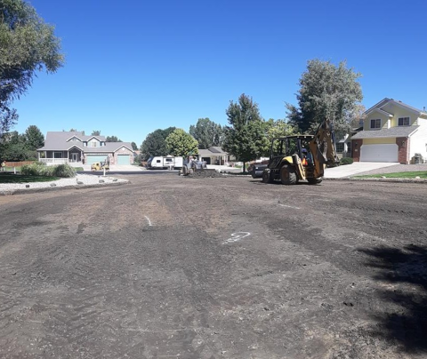 Photo of an improvement district road being prepared for paving. Workers are seen clearing debris and leveling the surface with machinery. Nearby houses and trees can be seen in the background.