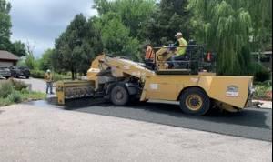 Photo of a road in an improvement district being paved with an asphalt paver machine. Surrounding houses and trees show in the background.
