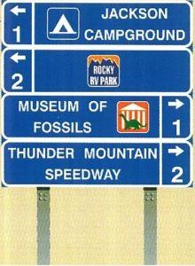 A Tourist Oriented Direction Sign (TOD) with four panels displaying location names, camping symbols, company logos, directional arrows, and distances. The panels provide information to help tourists navigate the area.