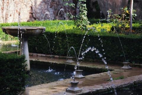 A brick-lined pond with several small fountains and a bird bath, surrounded by lush green hedges.
