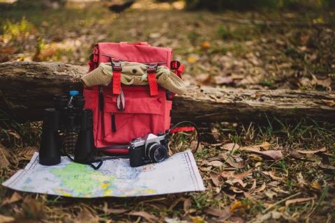 A red backpack, binoculars, a camera, and a map placed on a log in a hiking area with a field in the background.