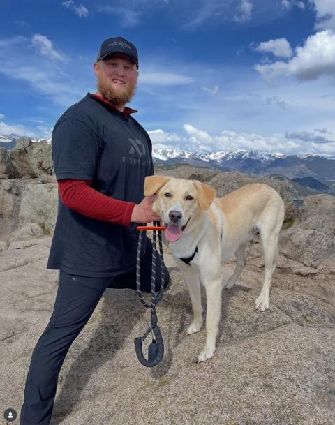 A man stands with a white dog on top of rocks with a view of the mountains