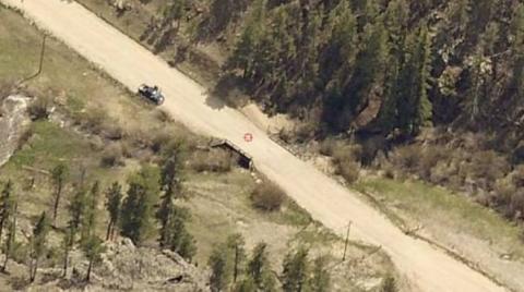 Aerial view of a dirt road passing through a forested area with a small bridge over a creek. A vehicle is parked on the roadside near the bridge. Surrounding the road are dense trees and sparse brush.