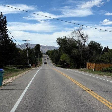 A view down Country Club Road, a two-lane rural road lined with trees and utility poles, stretching toward distant mountains. A wooden fence is on the right. A partly cloudy blue sky fills the background.