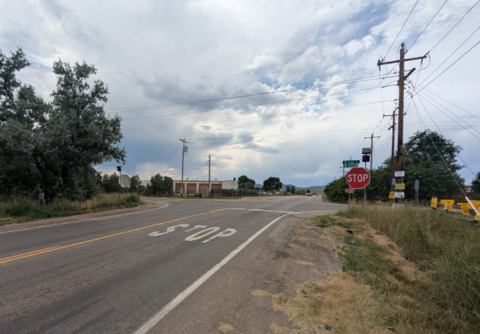 CR 70 & CR 15 intersection with a painted “STOP” marking on the pavement and a stop sign on the right side. Power lines and utility poles run along the roadside. Trees and grass line the edges of the road, and a few low buildings sit in the distance beneath a cloudy sky.