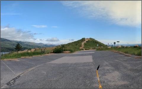 Paved parking overlook with empty spaces leading to a grassy hill and trail, with mountains and a valley visible under a partly cloudy blue sky; a yellow measuring rod lies in the foreground.