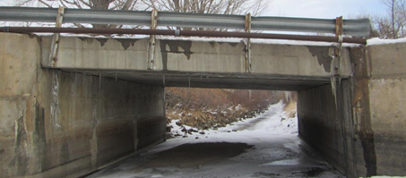 A small concrete bridge over a frozen, snow-covered creek. The bridge shows dark water staining and efflorescence on the fascia, with a rusted metal guardrail on top and small icicles hanging from the concrete deck.