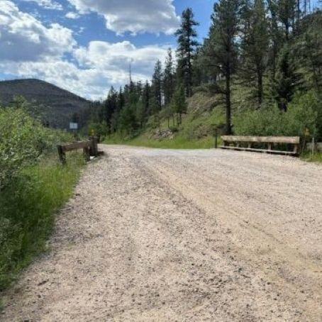 Unpaved dirt road crossing a small wooden bridge framed by pine trees and distant hills under a blue, partly cloudy sky.