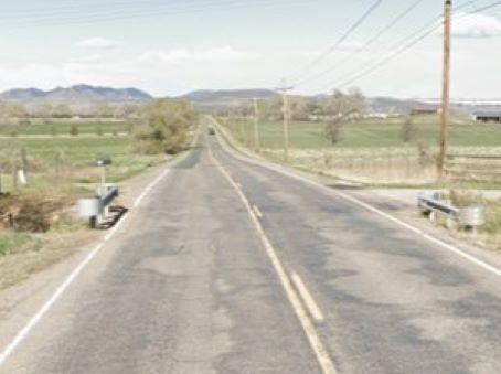 Paved two-lane rural section of Owl Canyon Road (CR 70) featuring patched asphalt, white edge lines, double yellow center lines, and metal guardrails on both shoulders. Utility poles, grassy fields, and mountain peaks frame the background.