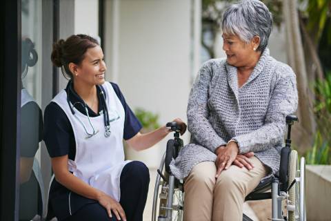 A medical professional kneels next to a smiling older adult in a wheelchair