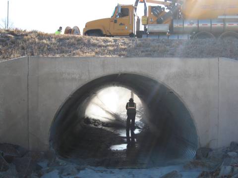 Larimer County employee power washing inside of culvert with a Larimer County truck on road above.
