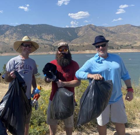 Три человека стоят у водохранилища Horsetooth с мешками для мусора после уборки мусора.