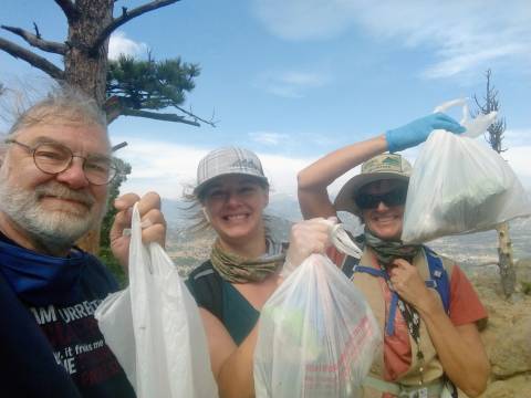 Three volunteers smiling at the camera and holding up the trash bags they filled while picking up litter at Hermit Park Open Space.