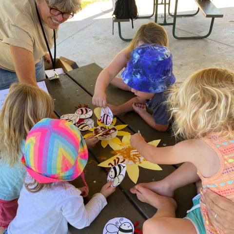 Volunteer assisting a group of preschoolers with bee craft at picnic table.