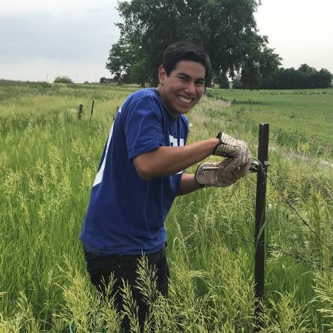 Man standing next to fence in a field of tall grass smiling at the camera and cutting barbed wire.