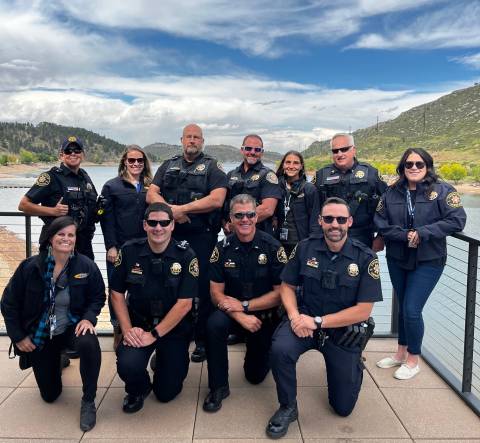 A group of smiling deputies and mental health clinicians stand in front of a large body of water with green hills in the background and a blue sky with white clouds overhead. 