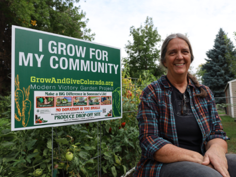 Een vrijwilliger van Master Gardener zit in haar tuin en glimlacht. Ze zit naast een Grow and Give-bord.