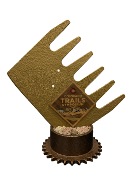 A close-up of the Golden McLeod Award trophy from the Colorado Trails Symposium, shaped like a trail tool with a cog base and filled with rocks.